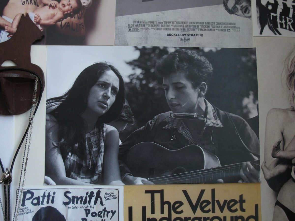 Black-and-white photograph of Joan Baez and Bob Dylan; Baez looks serious while Dylan plays an acoustic guitar with a harmonica around his neck. The photo is part of a collage that also includes posters for Patti Smith and The Velvet Underground.
